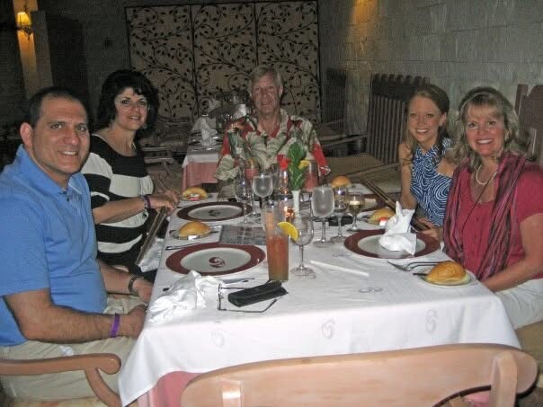 Six adults sit around a dining table in a restaurant, with plates, glasses, bread rolls, and a centerpiece.
