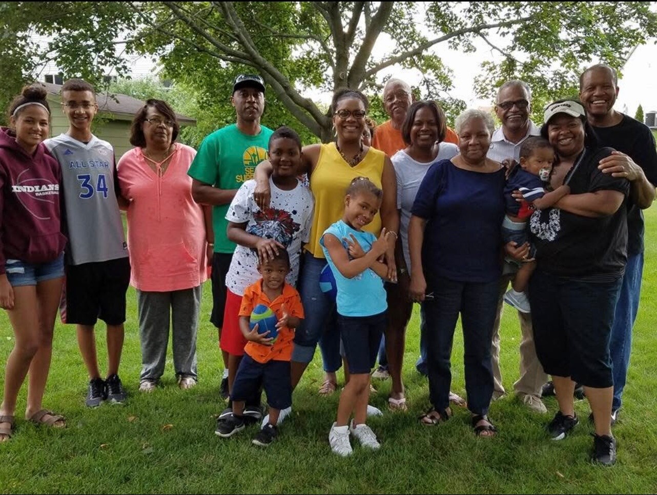 Group of diverse people, adults and children, posing together outdoors on a grassy field with trees, smiling at the camera.