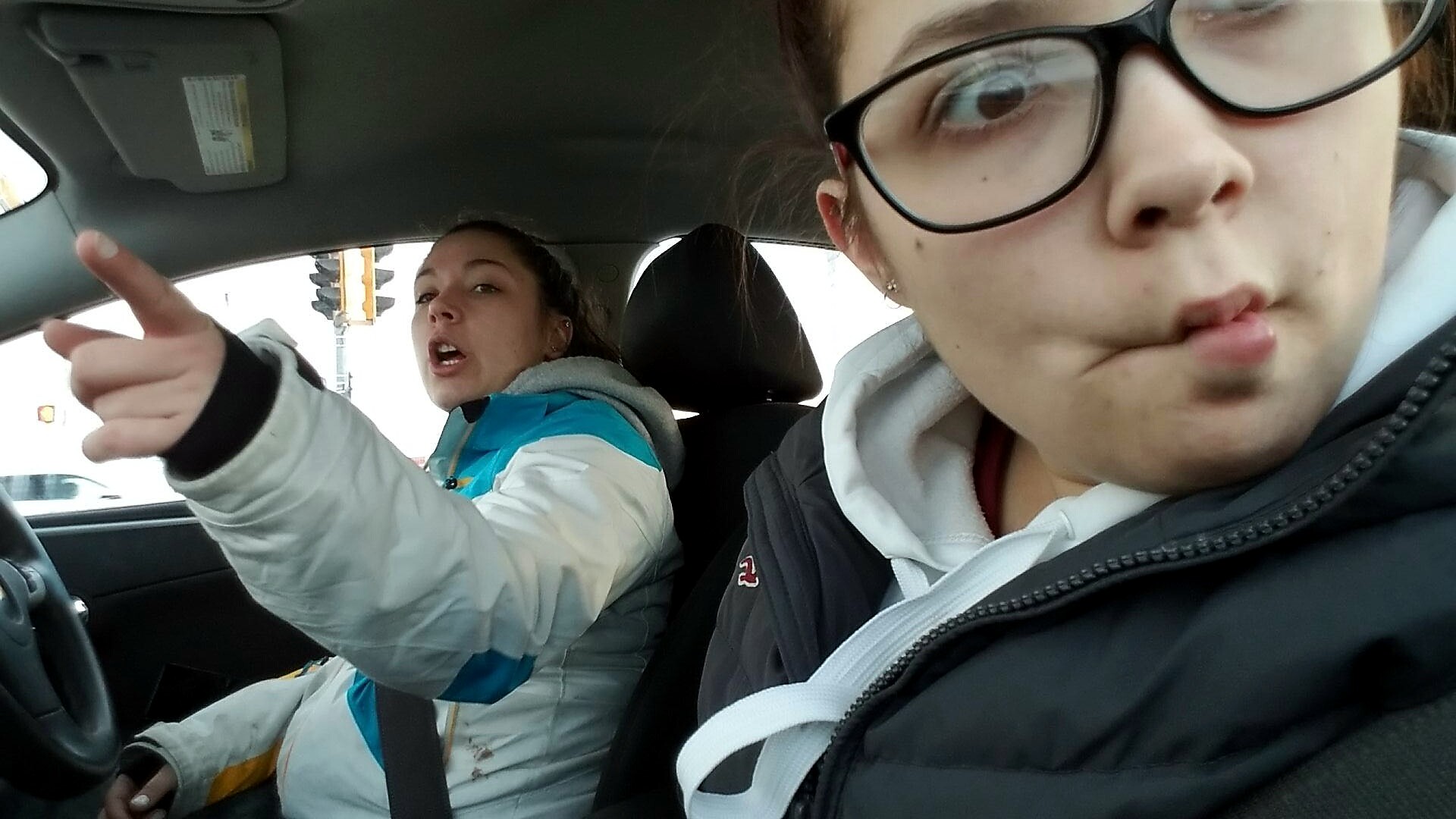 Two women in a car: driver in a blue-and-white jacket pointing ahead, passenger in the foreground wearing glasses making a puckered face.