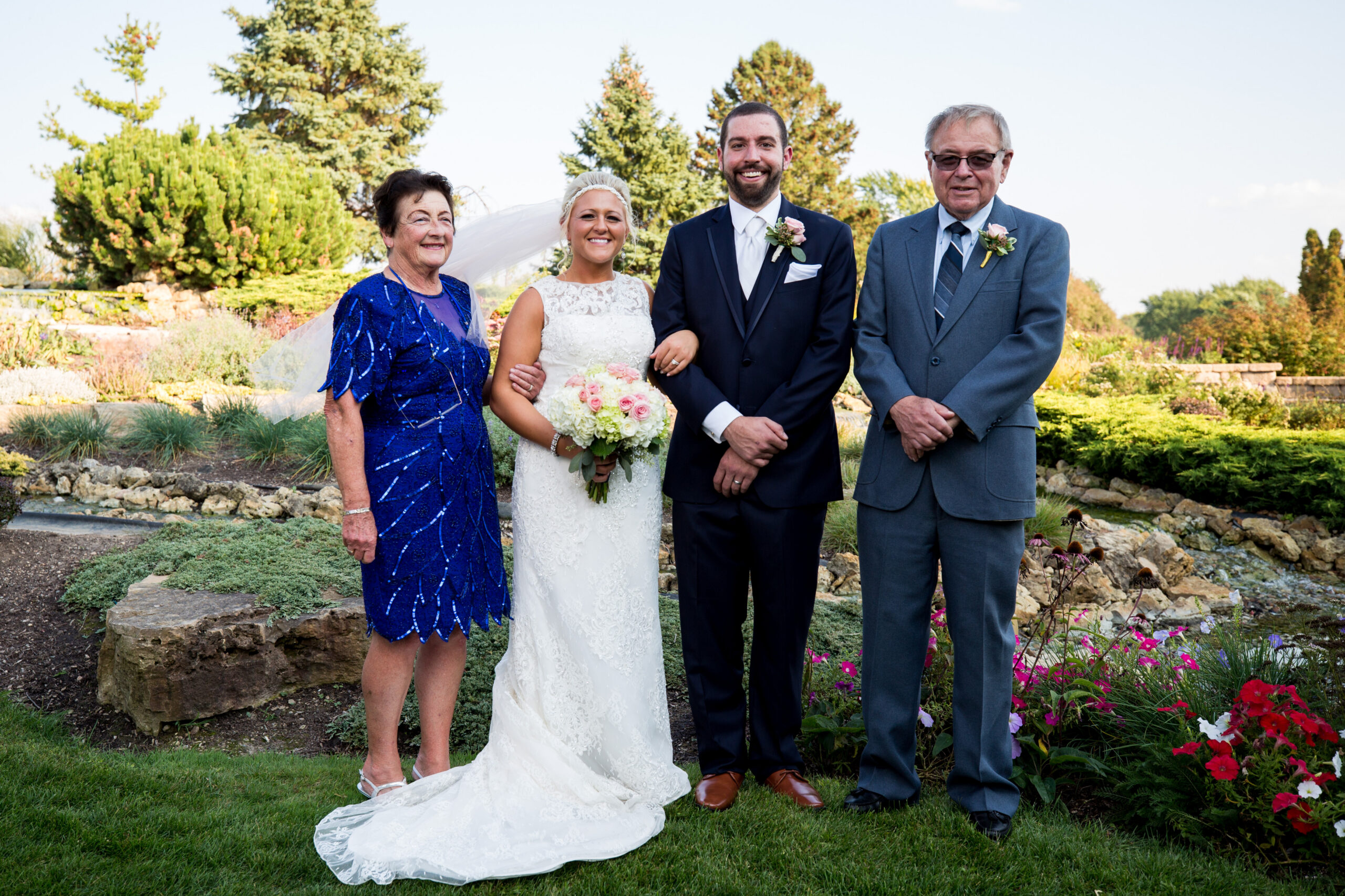 Bride in a lace white wedding dress holding a pink and white bouquet, flanked by a groom in a navy suit and two older guests in blue and gray outfits in a garden setting.