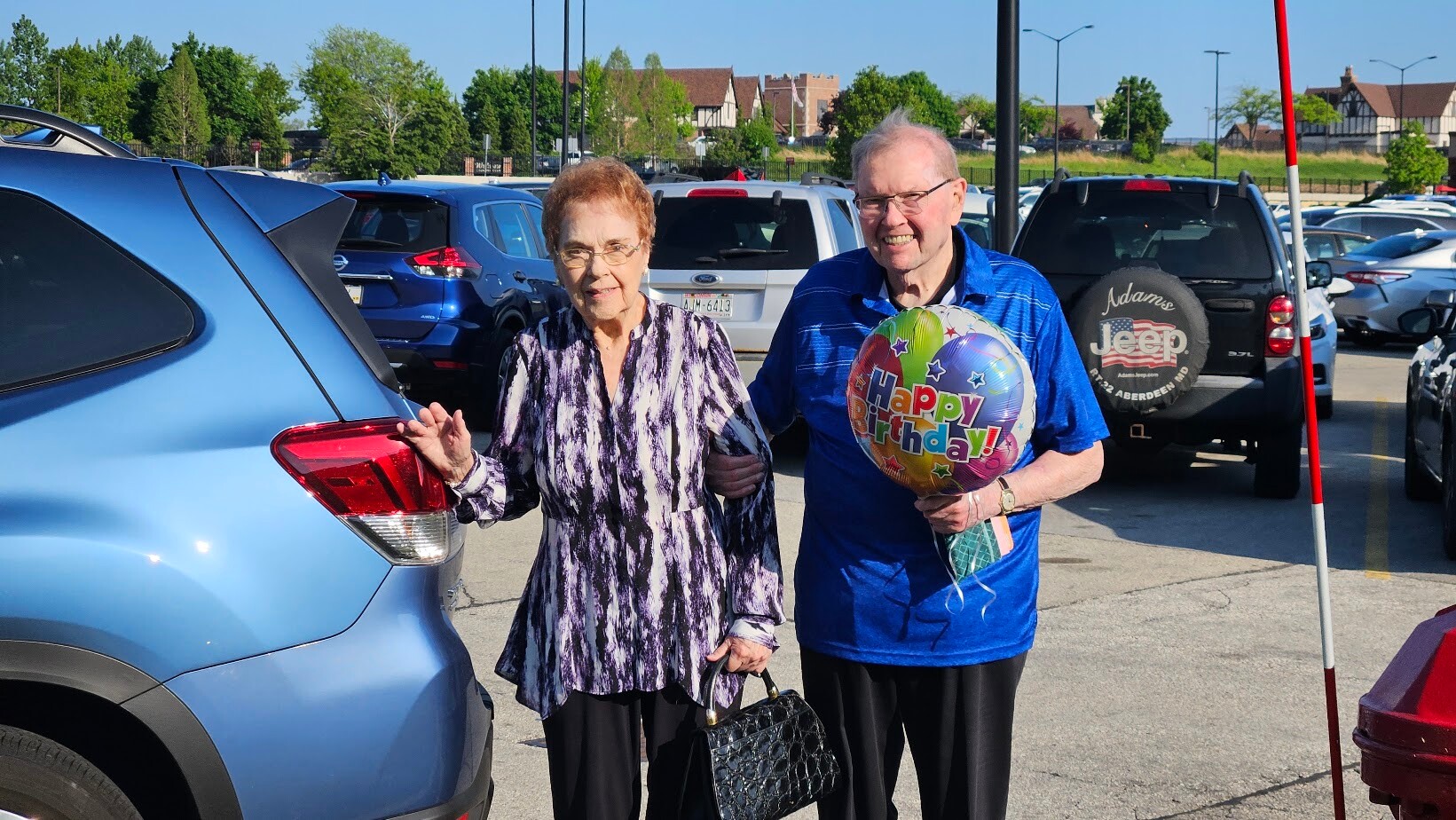 Smiling elderly couple stands in a parking lot beside a blue SUV; the man holds a colorful 'Happy Birthday' balloon.