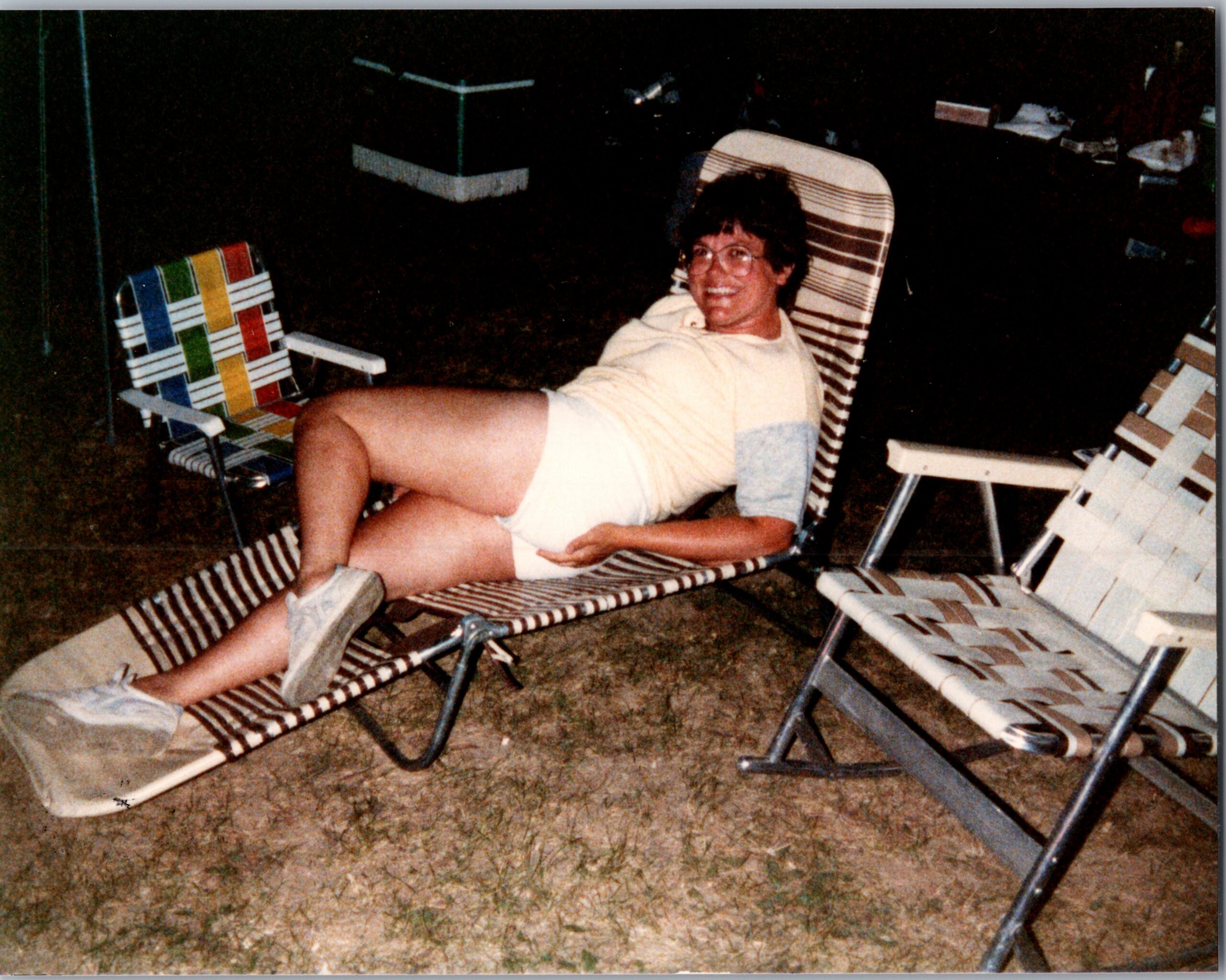 Man with glasses relaxing in a striped lawn chair in a backyard at night, surrounded by more lawn chairs.