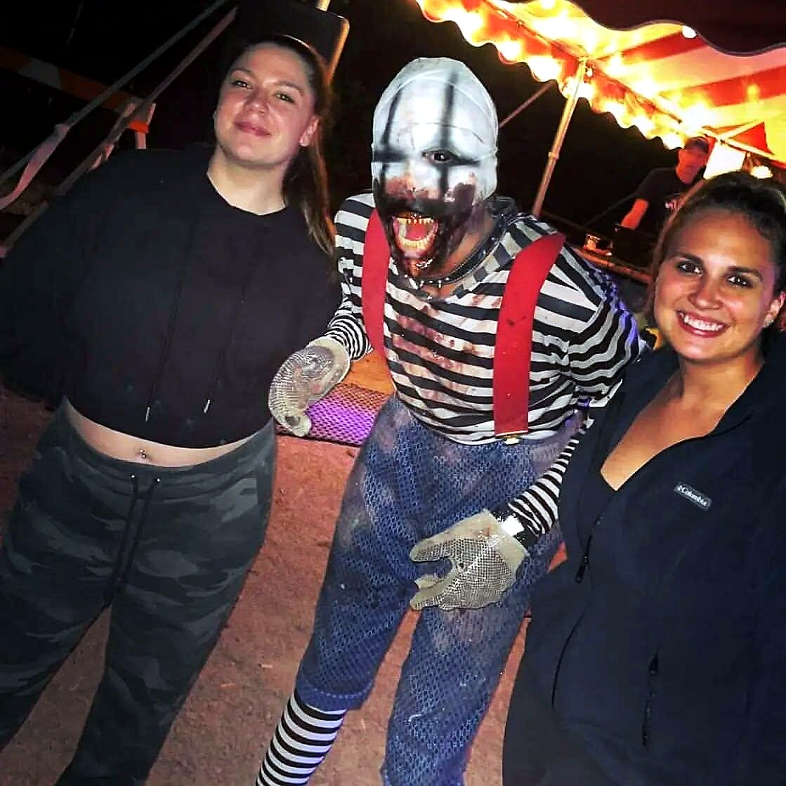Three people posing with a costumed mime in striped shirt, red suspenders, and metallic mask at a carnival.