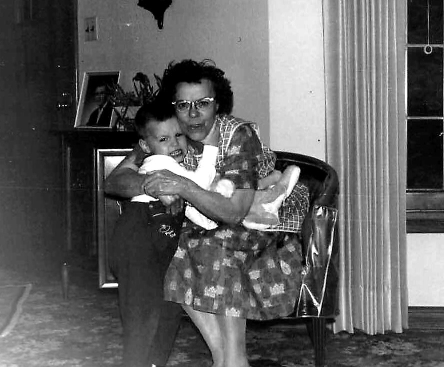 Older woman with glasses embracing a young child in a living room, both looking at the camera.