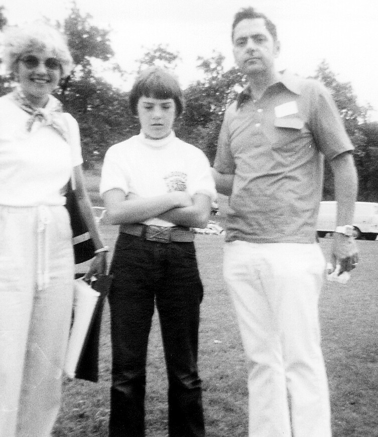 Three people stand outdoors on a grassy area: left a smiling woman with light hair, center a person with arms crossed, right a man in a short-sleeve shirt and light pants.