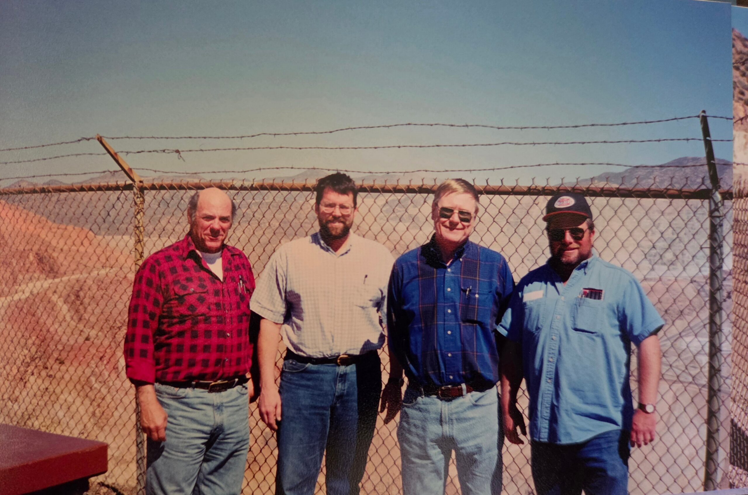 Four men posing in front of a chain-link fence with barbed wire, desert landscape and distant mountains under a clear blue sky.