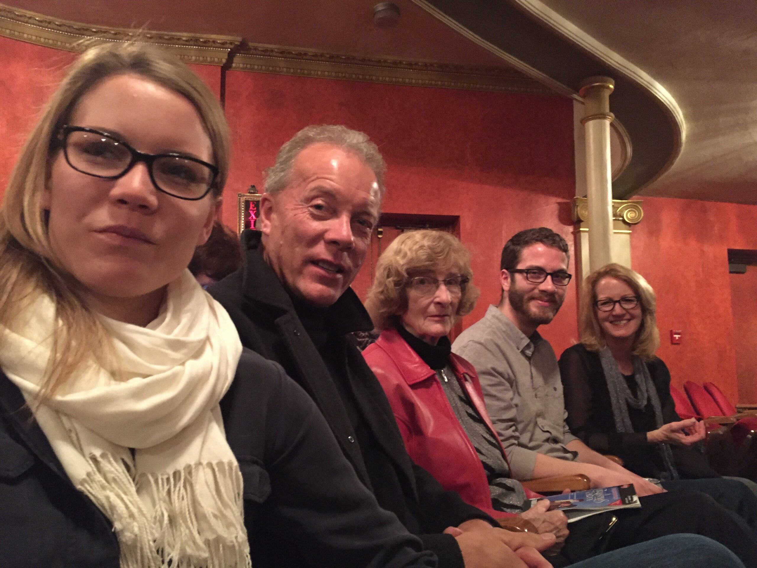 Close-up selfie of several people seated in a theater with red walls, smiling at the camera (front-left woman with glasses and scarf).