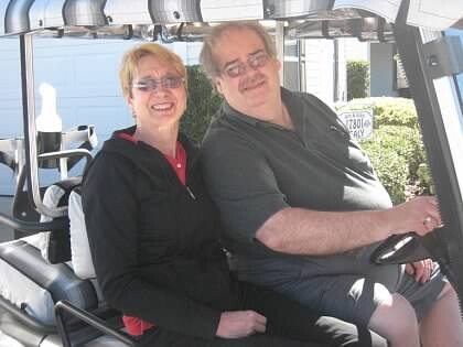 Smiling couple seated in a golf cart; woman with short blonde hair and glasses on the left, man with gray hair on the right, outdoors on a sunny day.