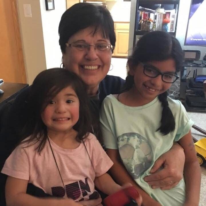 A woman hugs two young girls indoors, all smiling at the camera.