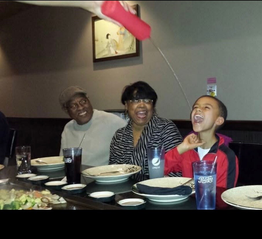 Family of three at a restaurant table laughing as a pink drink is poured into the boy's mouth from above.