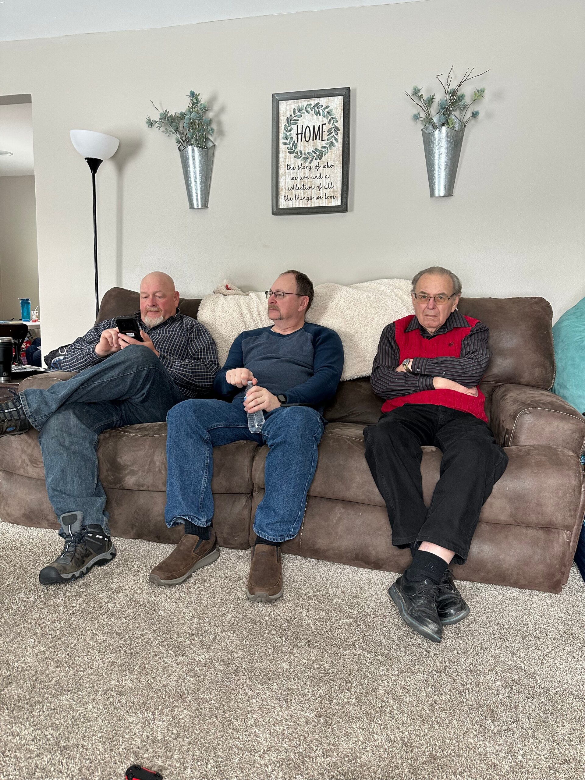 Three men sit on a brown couch in a living room; the left man uses a smartphone while the other two gaze forward.