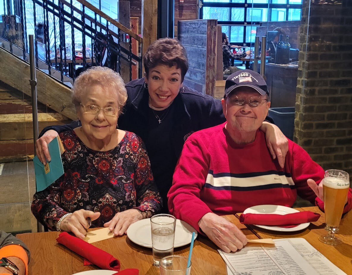 Three smiling friends seated at a wooden restaurant table: an elderly woman on the left holding a turquoise card, a woman in the middle leaning in, and a man in a red sweater and cap on the right with a beer glass nearby.