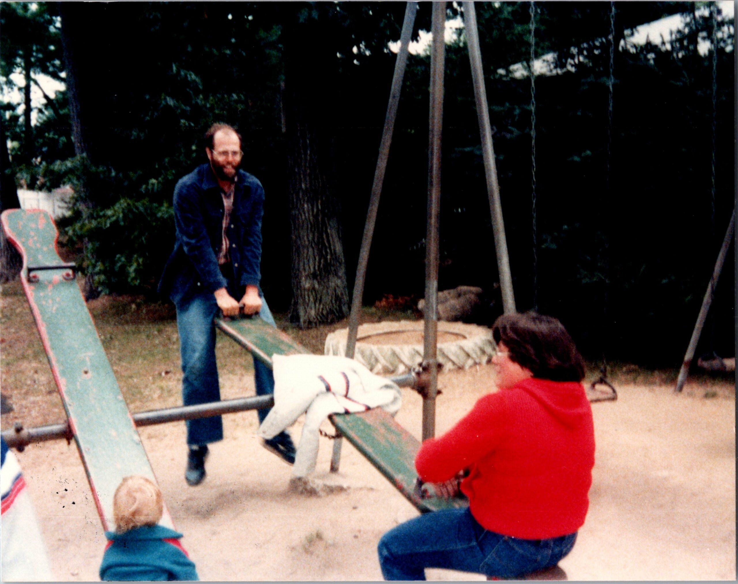 Man with a beard in a denim jacket standing by a wooden swing set as children play on the swings in a backyard park with trees in the background.