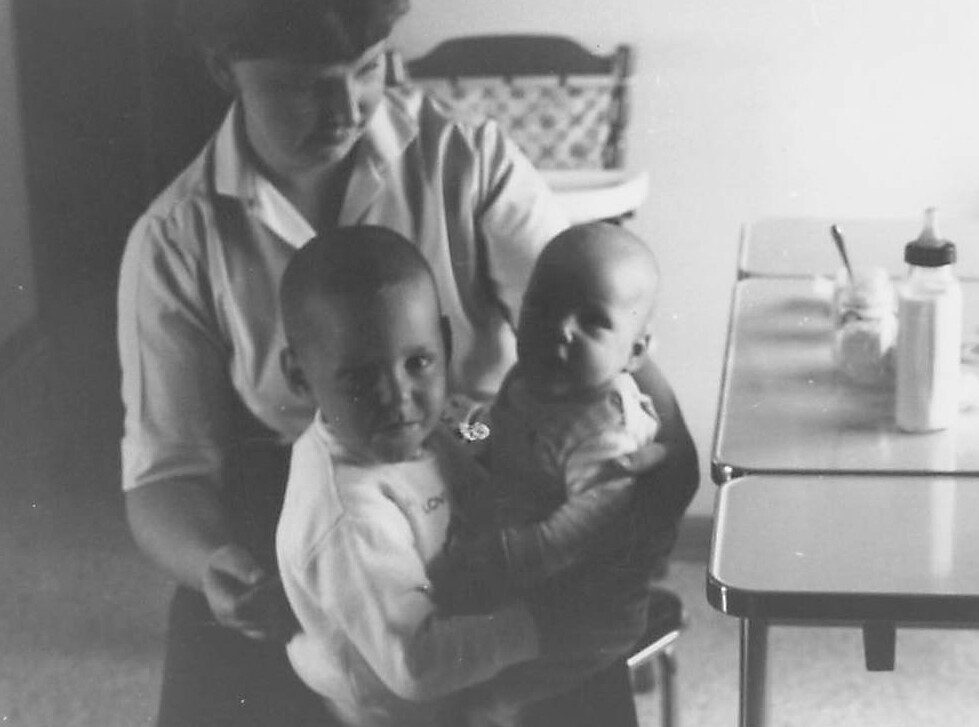 Woman standing behind two young children, holding the younger baby as the older child faces the camera; a bottle sits on a nearby table.
