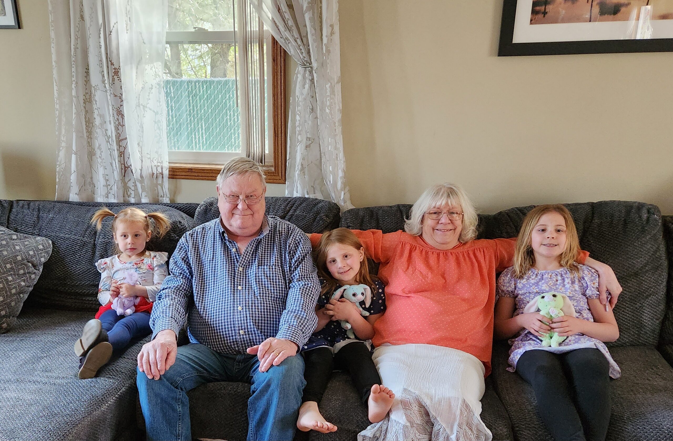 Family sits together on a dark sofa in a living room for a group photo, with two adults and three or four children holding stuffed animals and smiling at the camera.