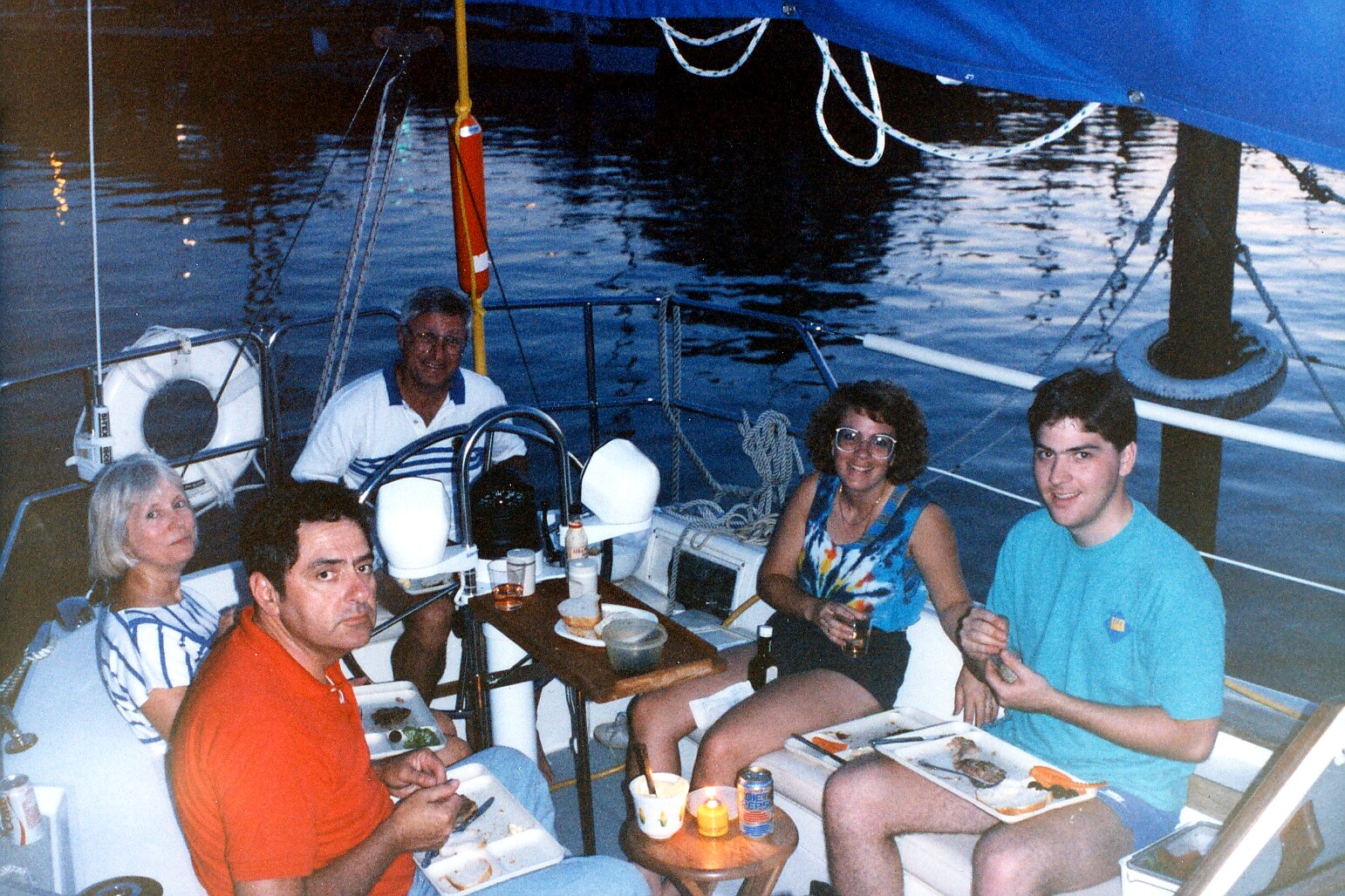 Group of five adults dining on a sailboat at night, seated around a small table with plates, drinks, and a candle, with blue canopy and water in the background.