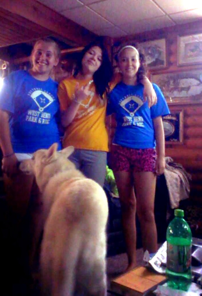 Three friends posing indoors in a wood-paneled room; two wear blue shirts, one in orange, with a dog in the foreground.