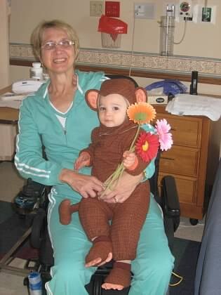 Woman in teal scrubs sitting in a hospital room with a baby in a brown monkey costume holding colorful flowers.