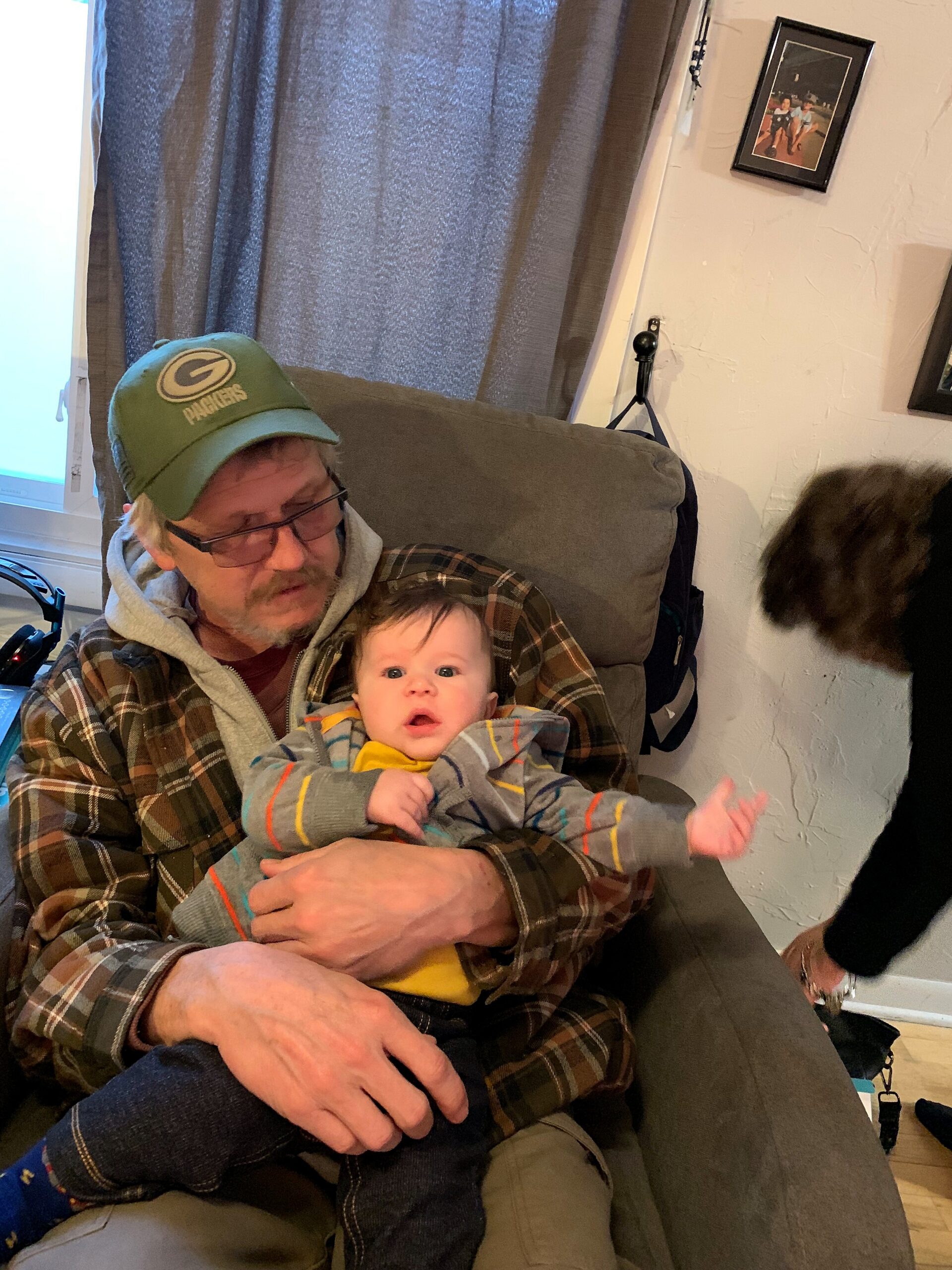 Man in a green Packers cap sitting in a chair, holding a baby wrapped in a striped onesie indoors.