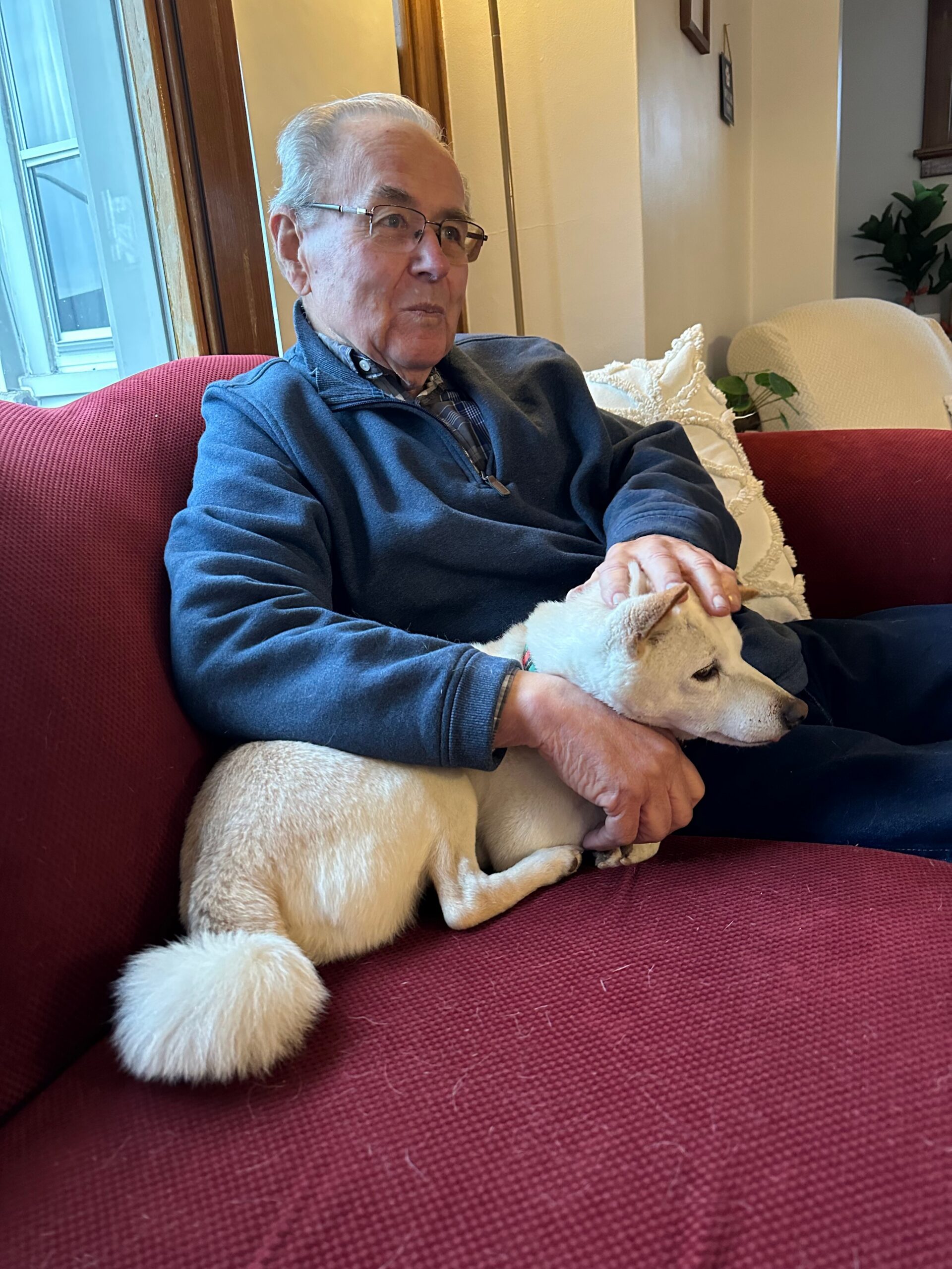 Older man wearing glasses sits on a red couch, gently holding a light-colored dog in his lap indoors.