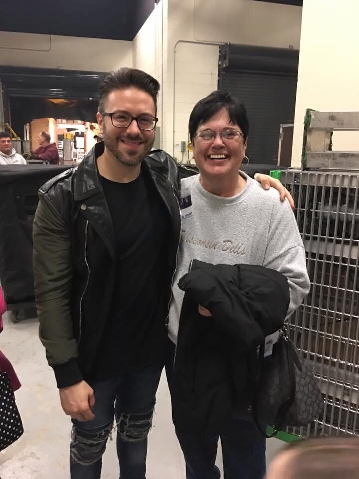 Two people pose backstage for a photo: man in a black jacket and glasses, woman in a gray sweatshirt smiling with a coat in her arms.