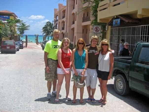 Six people posing together on a sunny seaside street with palm trees, the beach and ocean visible in the background.