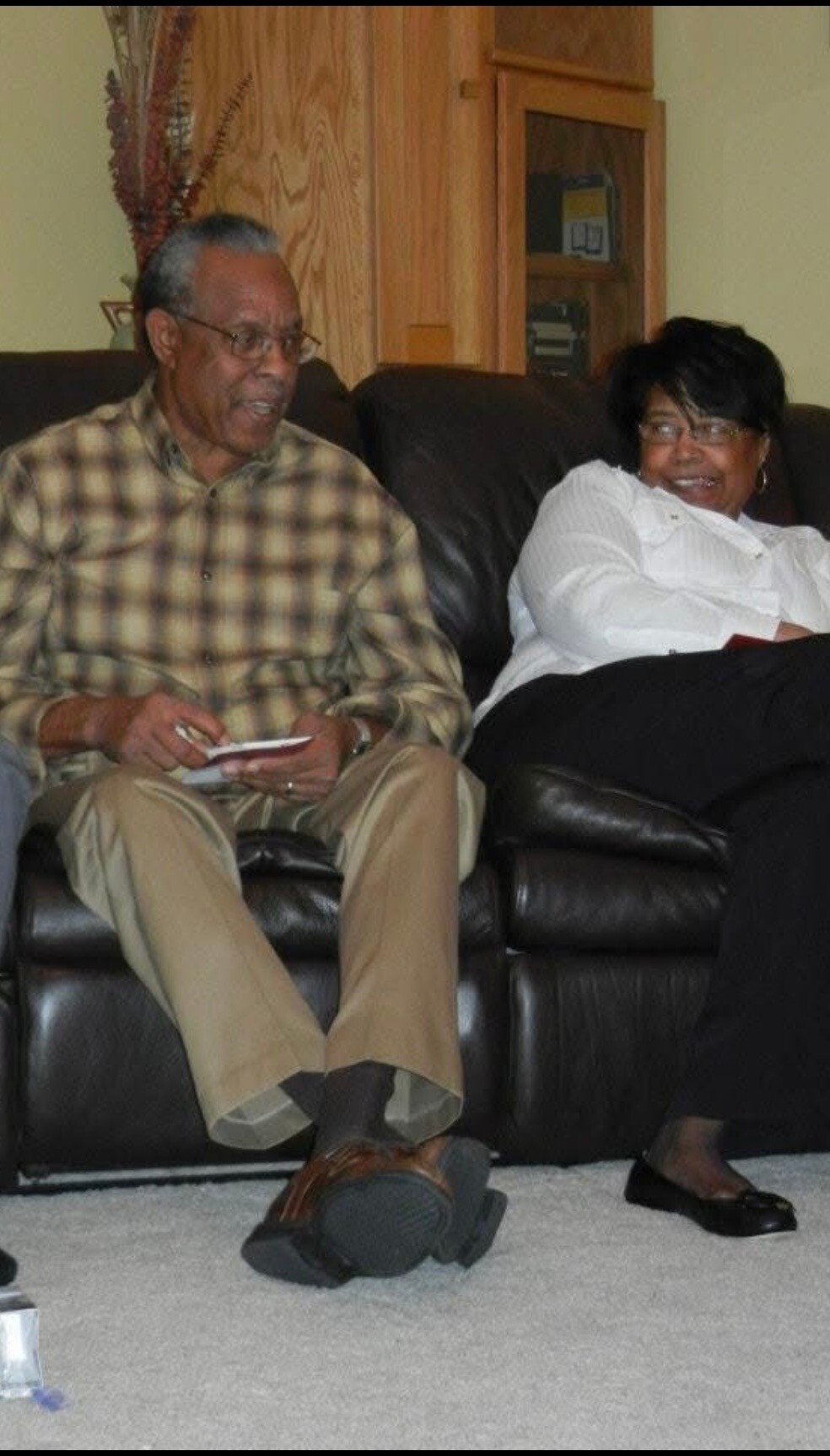 Two seniors sit on a dark leather sofa in a living room, smiling and chatting; the man wears a plaid shirt and khaki pants, holding a napkin.