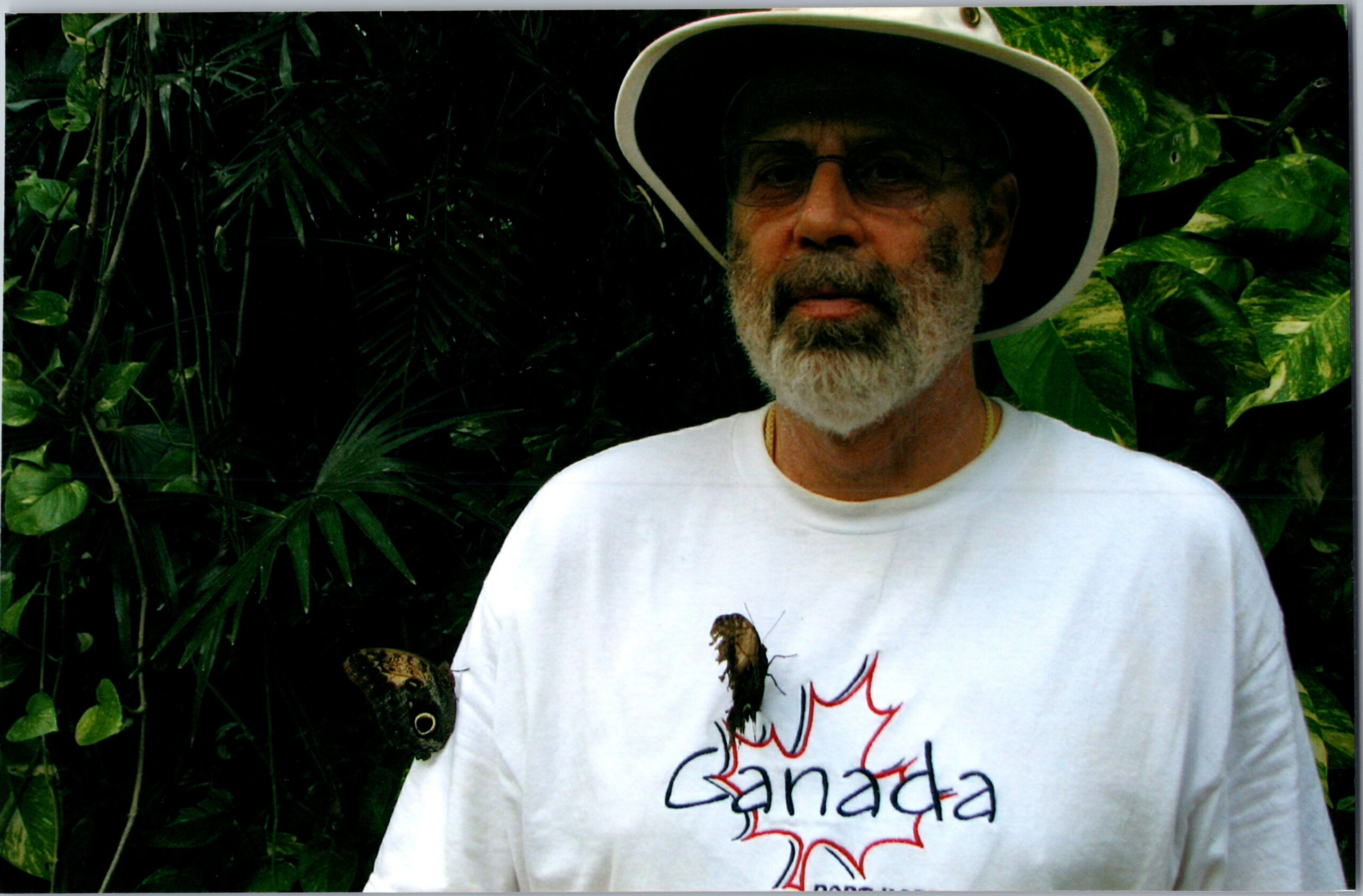 Older man with a white beard and wide-brim hat stands among tropical plants; white shirt shows a red maple leaf and the word Canada.