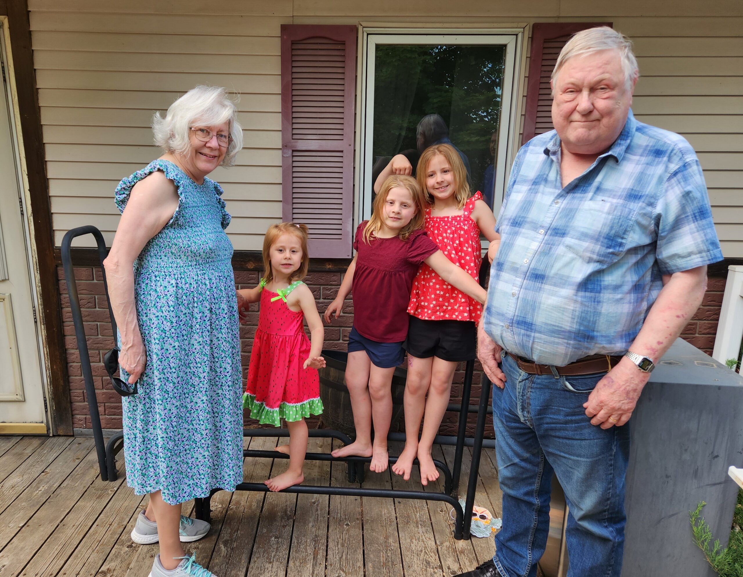 Grandparents with three young girls posing on a wooden porch in front of a beige house, all smiling at the camera.
