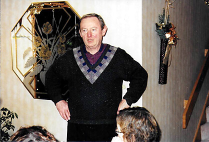 Man in a black sweater with a gray-purple V-pattern stands with hands on hips in a decorated living room with holiday decor in the background.