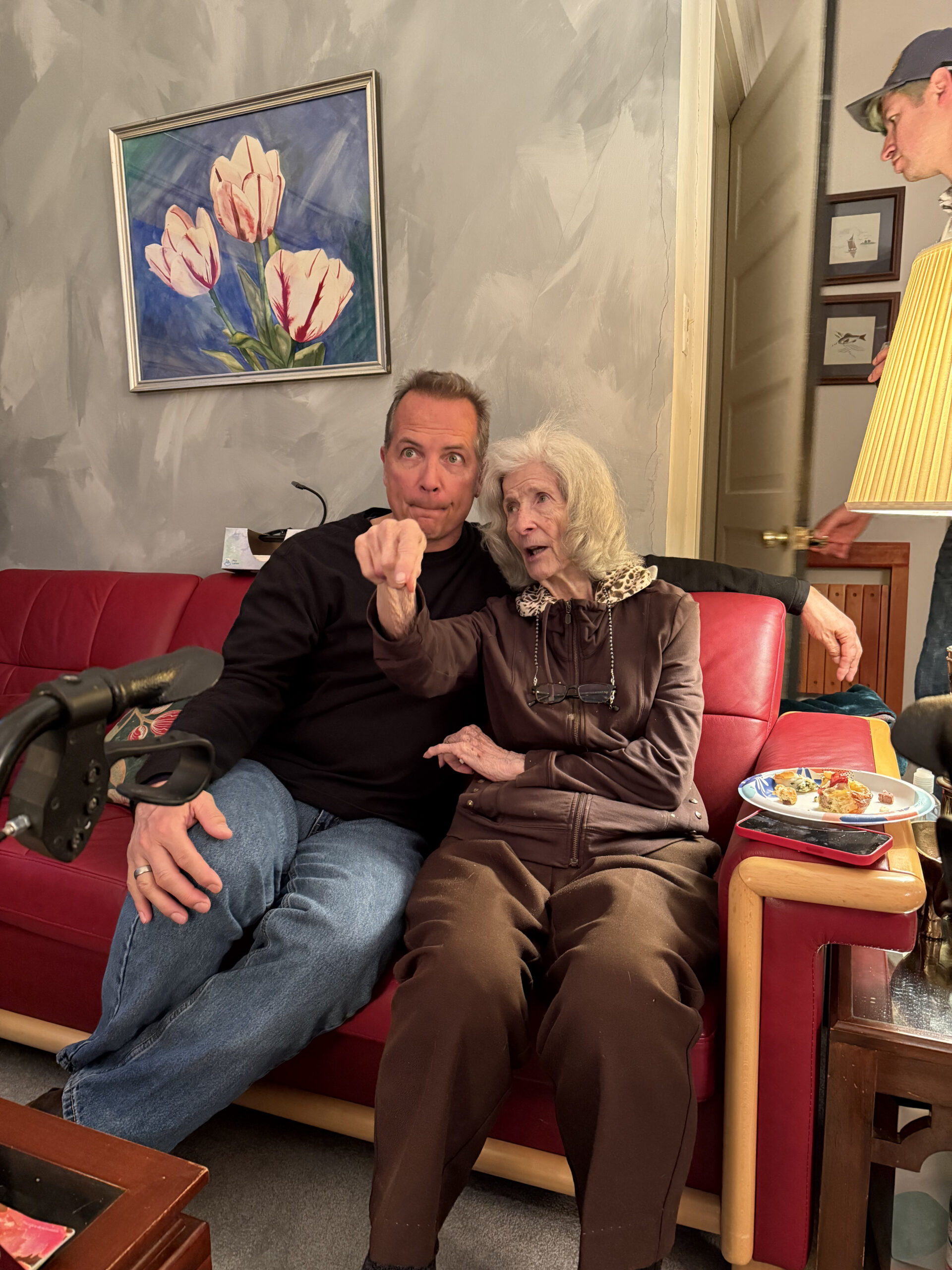 Two people sit on a red sofa in a living room: a man in a black shirt points toward the camera while an elderly woman beside him smiles, with a walker visible on the left.