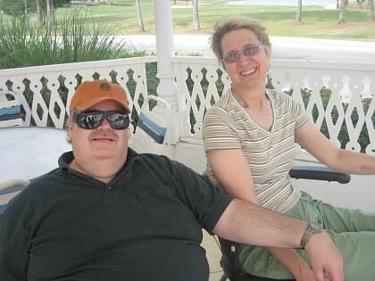 Two people seated on a sunny porch with decorative white railing; man wearing an orange cap and sunglasses, woman in a striped shirt.
