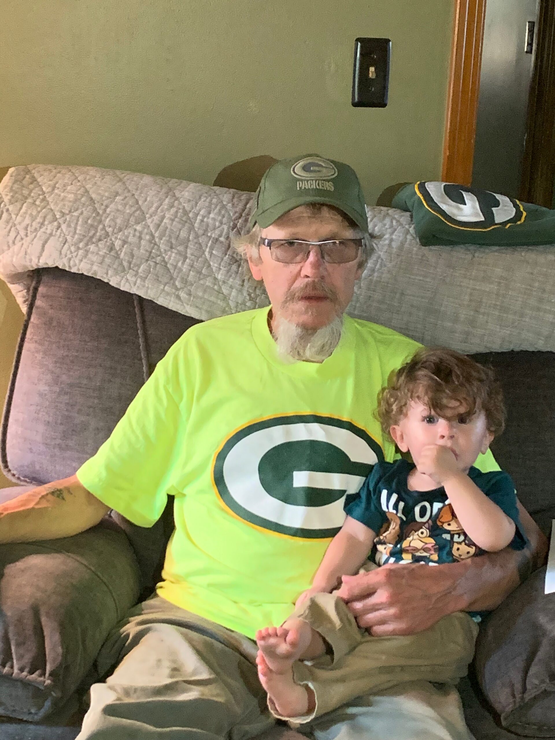 Older man with glasses and a gray beard wearing a Green Bay Packers cap and a neon green shirt sits on a sofa, holding a young child with curly hair on his lap.