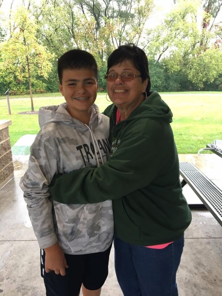Smiling boy in a gray hoodie and braces hugging an older woman in a green jacket at a park pavilion with trees in the background.