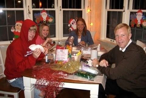 Five people sit around a table covered with craft supplies, making holiday decorations in a festively decorated room with Santa decals on the windows.