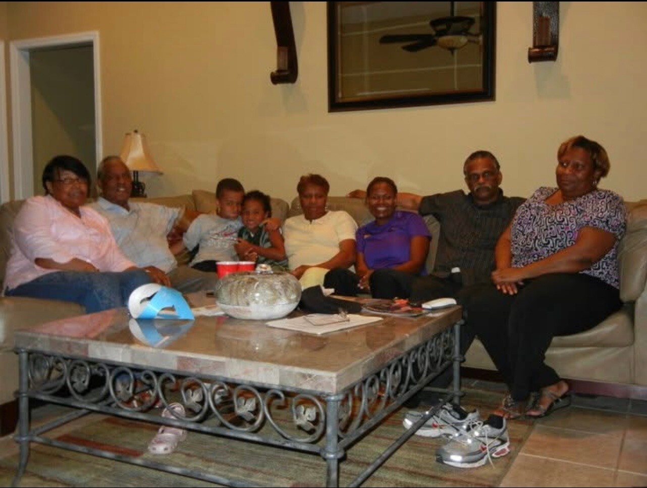 A multi-generational family sits together on a beige sofa in a living room, smiling at the camera with a coffee table in front.