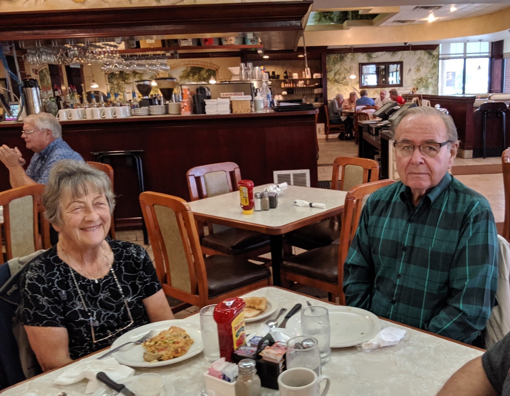 Smiling elderly woman and man sit at a restaurant table with pizza slices and condiments, bar area in the background.