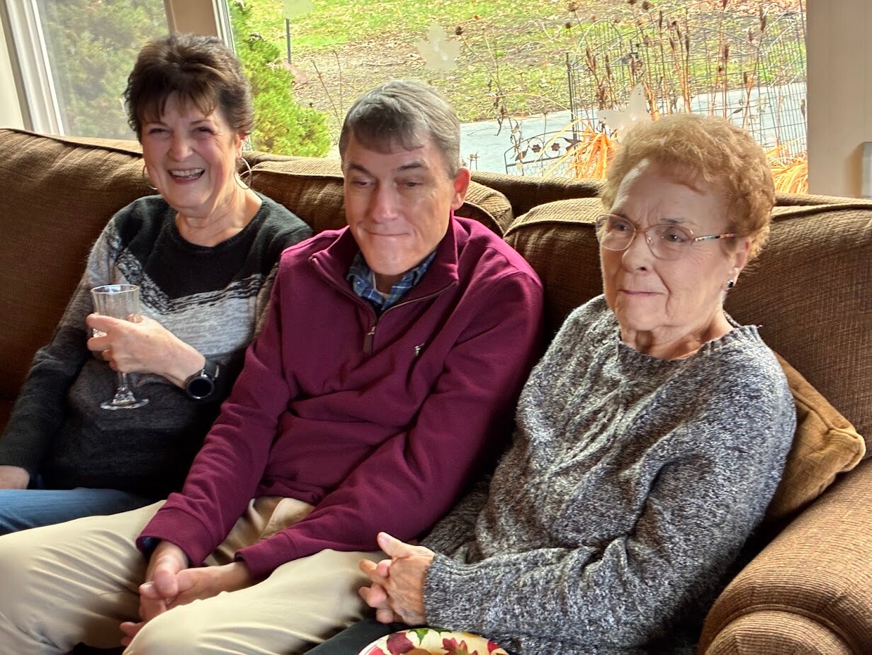 Three smiling seniors seated on a brown couch indoors; the woman on the left holds a champagne glass.