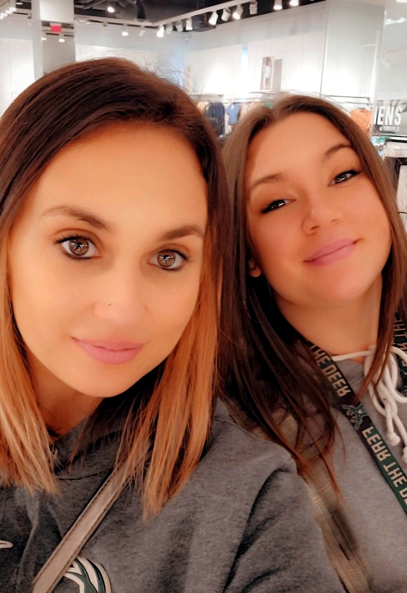 Two women smiling for a selfie in a clothing store with racks and bright lights in the background.