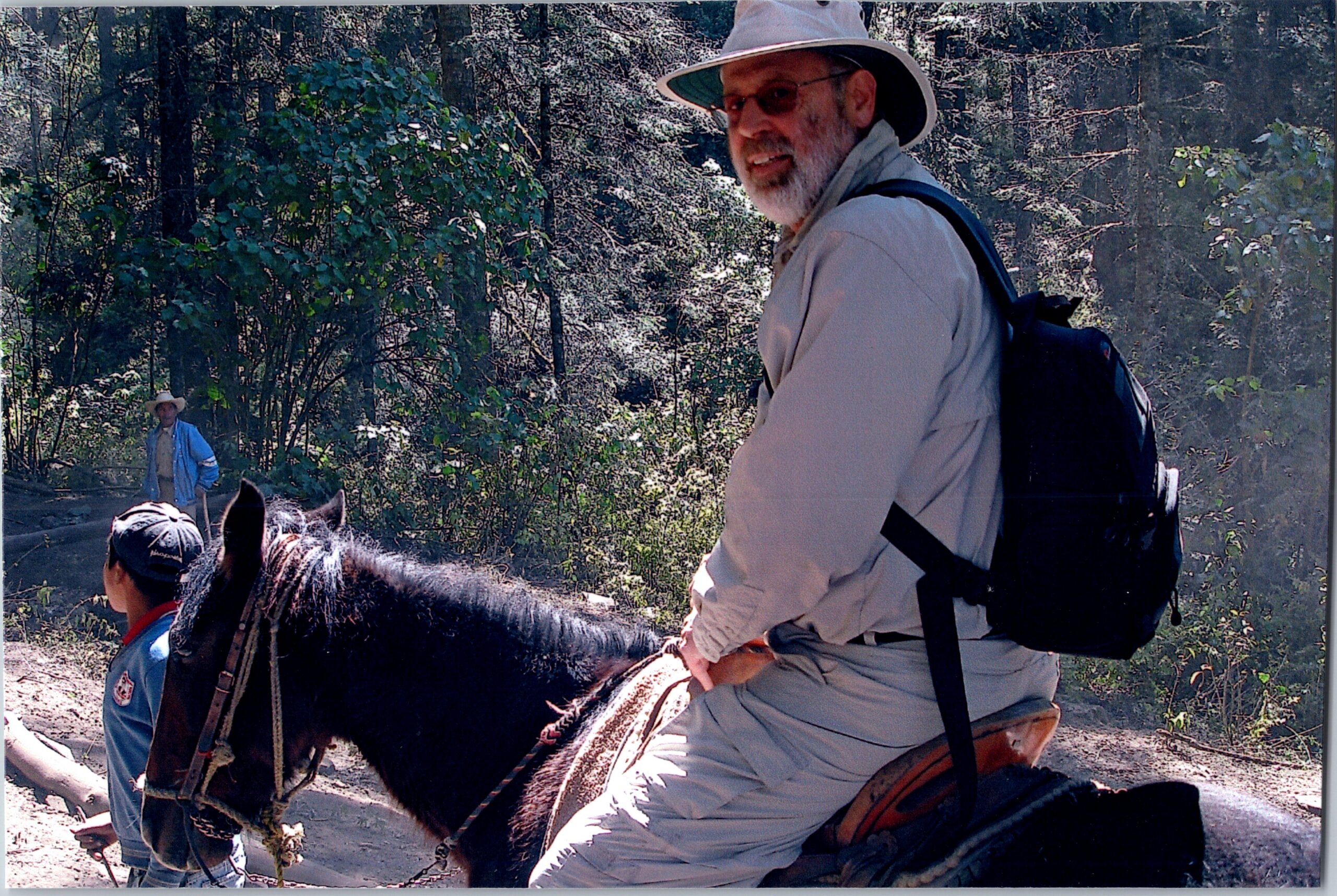 Bearded man in a wide-brim hat and backpack rides a horse on a forest trail; another rider is visible in the background.