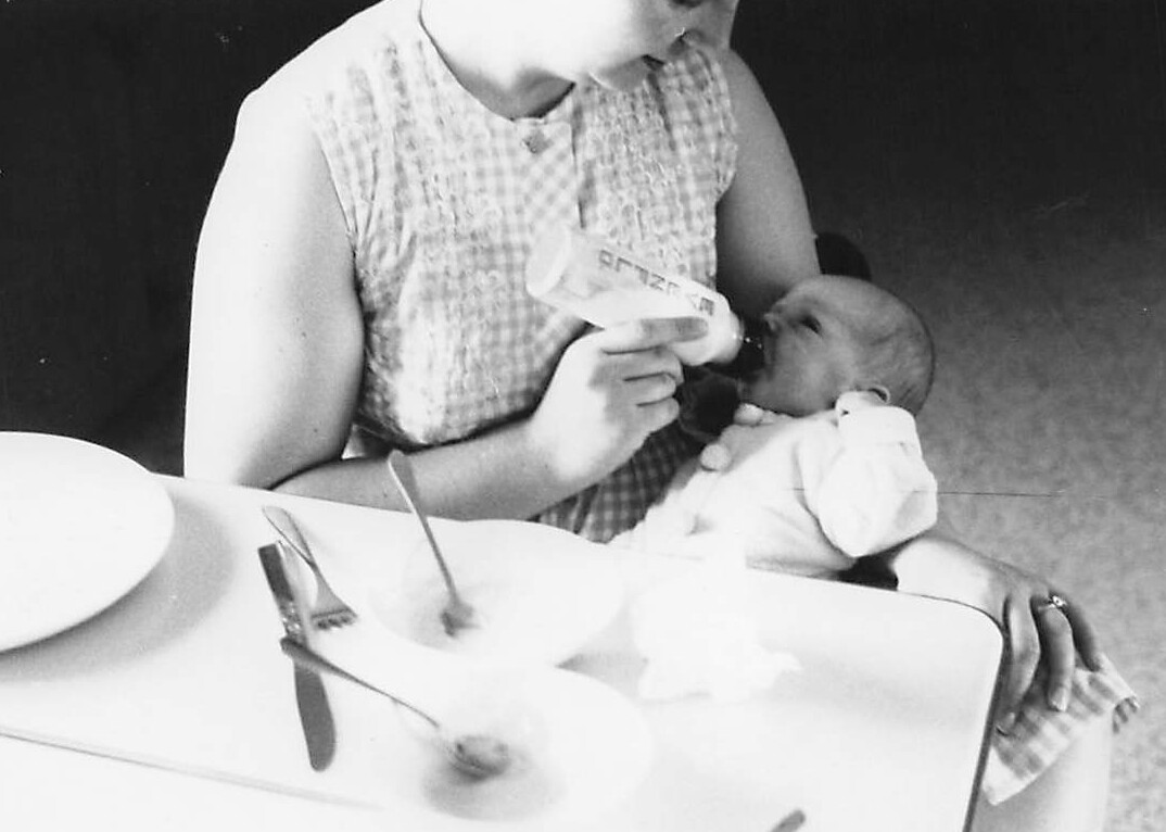 Woman bottle-feeding a baby held in her lap at a table.