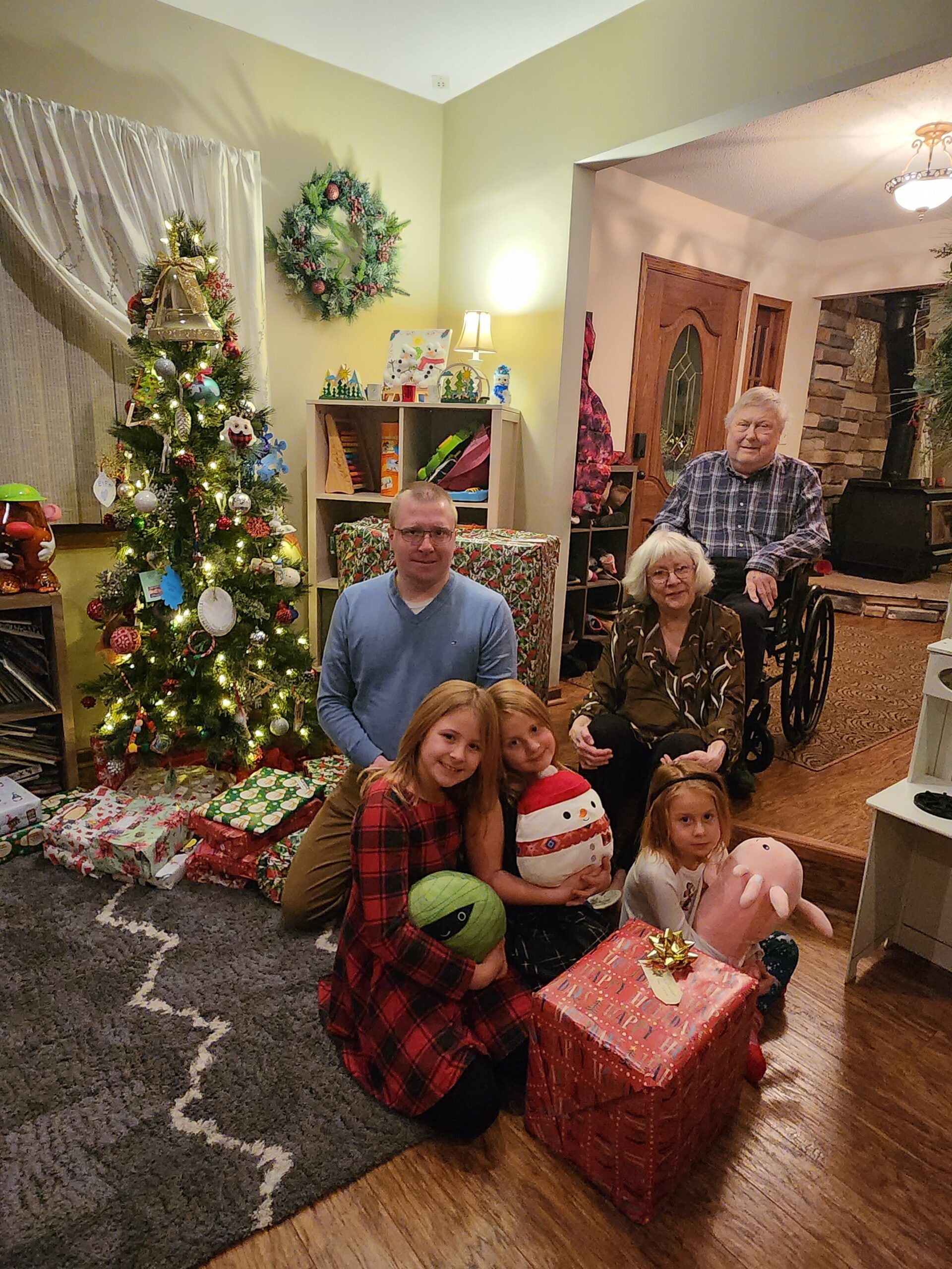 Family posing for a Christmas photo in a cozy living room with a decorated Christmas tree and wrapped gifts nearby.