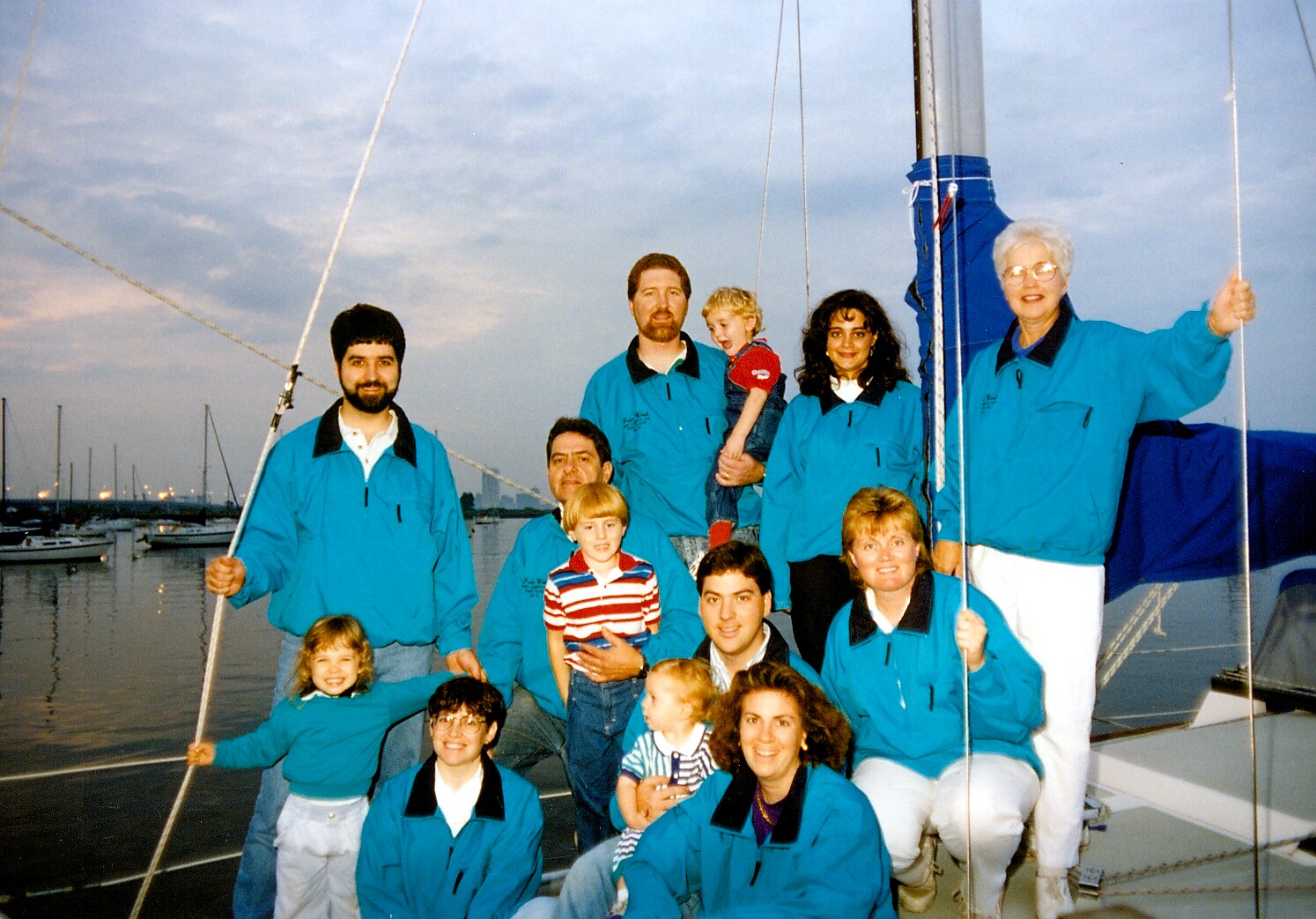 Group of people in matching teal jackets posing on a sailboat at dock, smiling for the photo, evening sky in background
