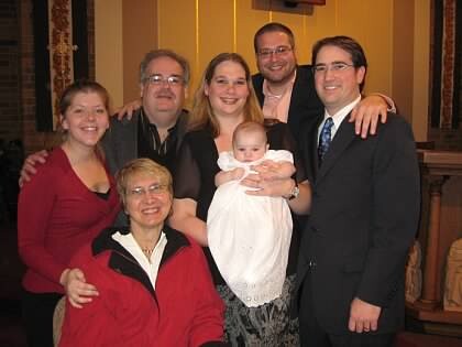 Group portrait of eight family members with a baby in a white dress at center wearing a lace-trimmed dress, smiling in a warmly lit room.