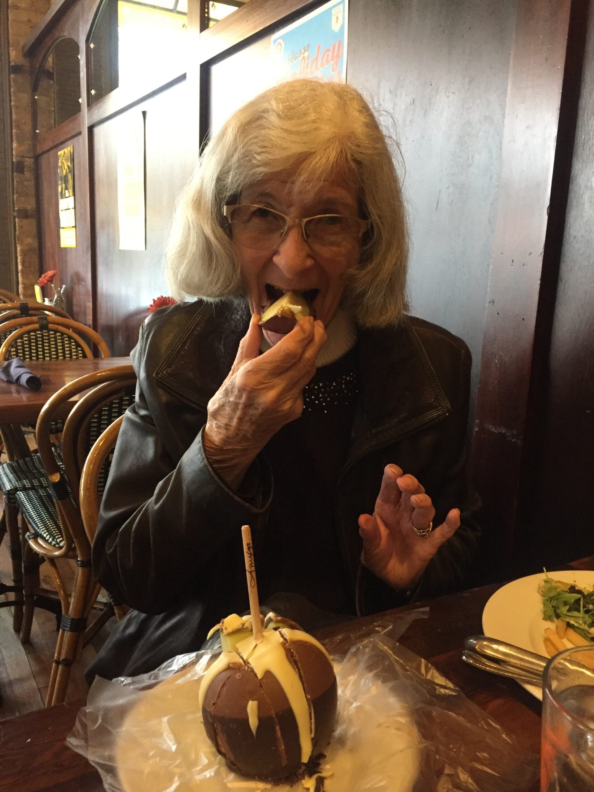 Elderly woman with gray hair and glasses takes a bite of a chocolate-dipped apple dessert at a restaurant.