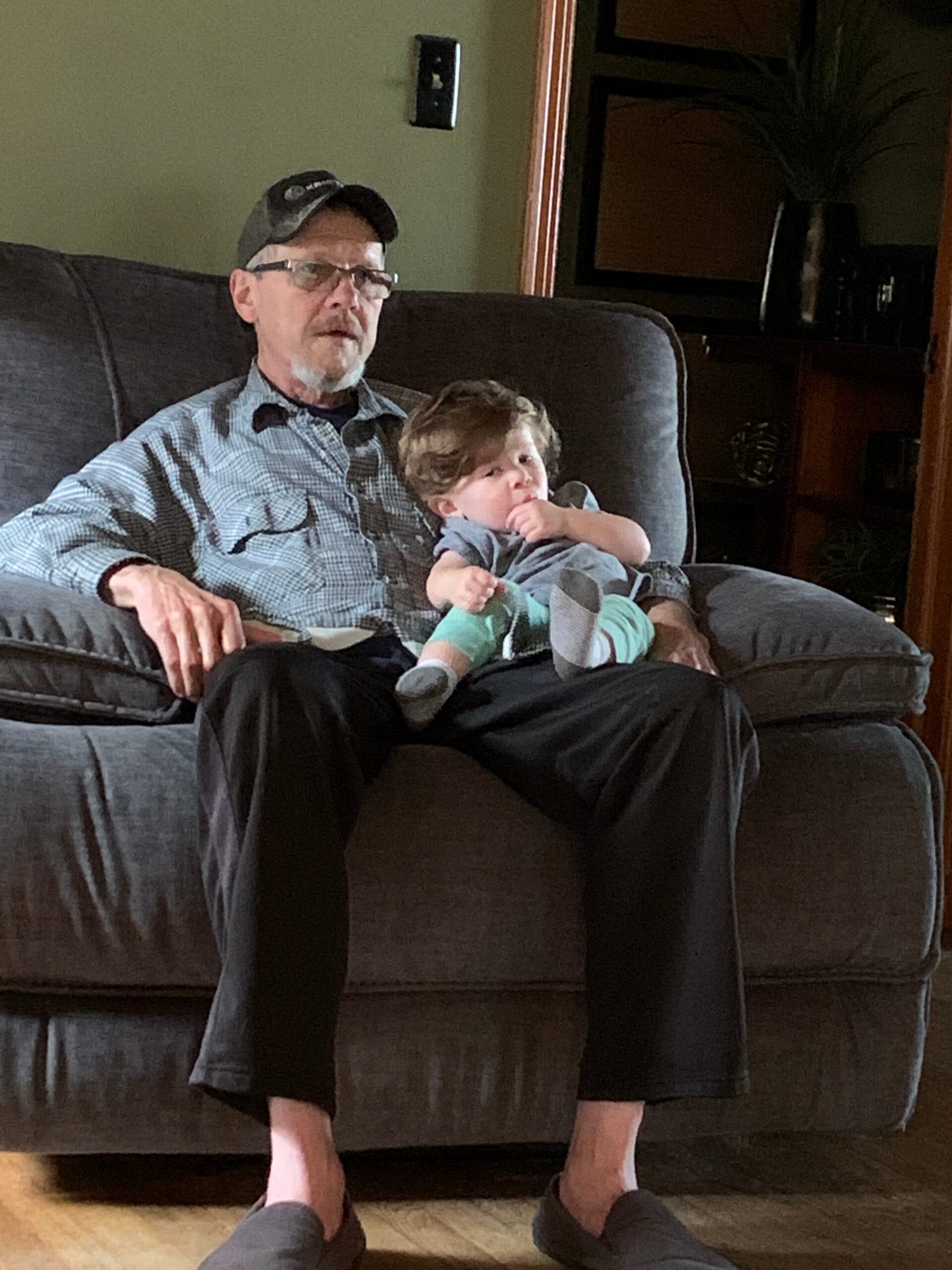 Older man wearing a cap and glasses sits on a gray sofa with a small child on his lap in a living room moment.