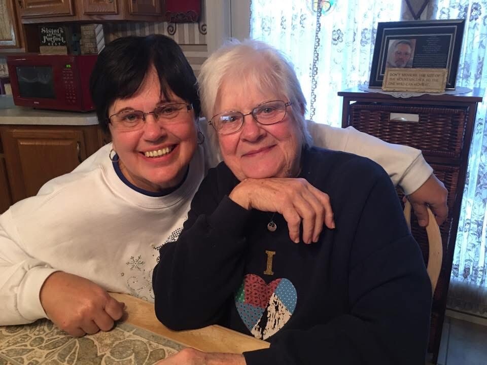 Two women sit at a kitchen table, smiling with their arms around each other in a warm family moment.