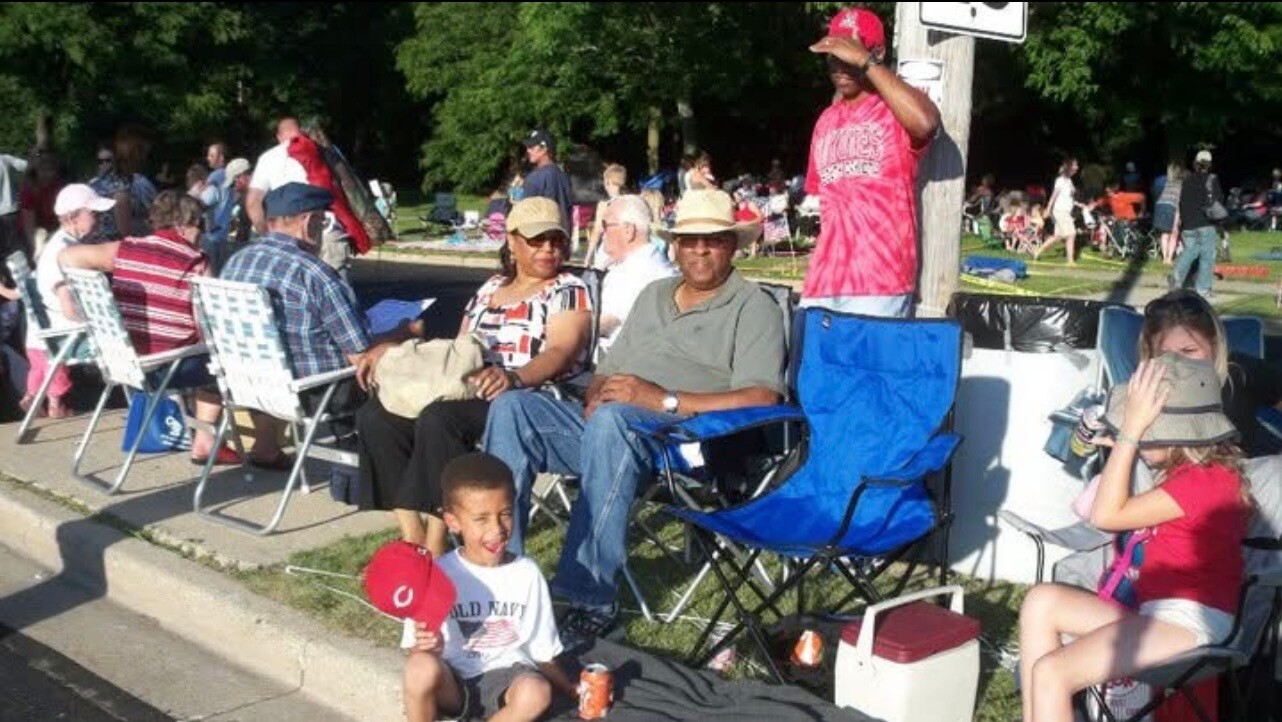 People sit in lawn chairs along a sidewalk at an outdoor event or parade; a boy in front holds a red cap and smiles while others chat in shade.