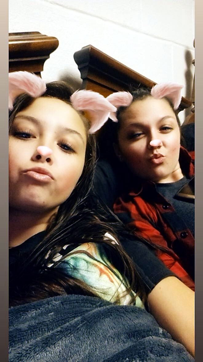 Two young women taking a selfie with pink animal-ear filter, posing with puckered lips in a cozy setting.