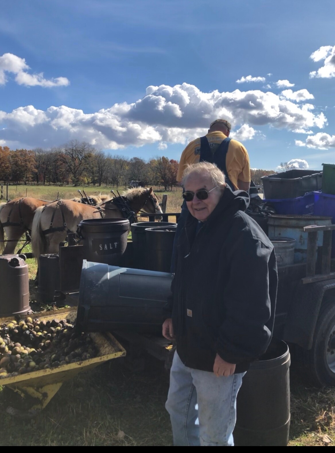 Elderly man wearing sunglasses and a dark jacket stands beside a wagon loaded with crates and barrels on a farm, with horses in the background.